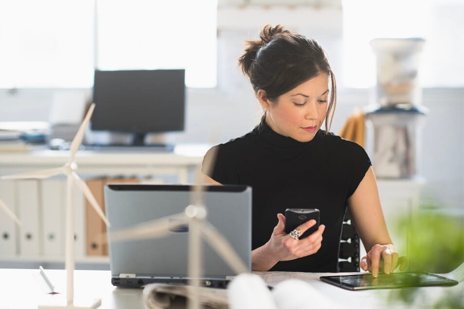 woman holding phone and tapping on tablet sitting in front of computer