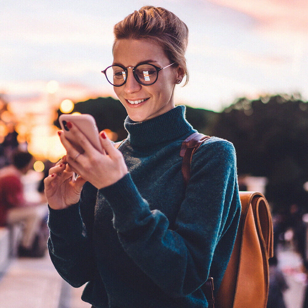woman in glasses and turtleneck looking at a phone outside